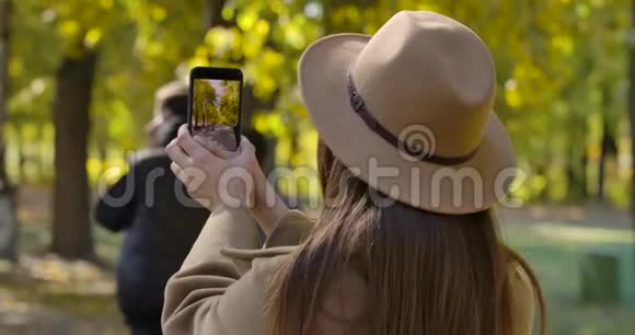 年轻的高加索女孩戴着棕色帽子在她的智能手机上拍摄秋天公园的照片愉快的欧洲视频的预览图