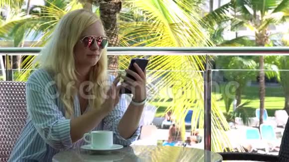 自由职业漂亮的金发女人在热带国家的咖啡馆里用智能手机工作喝咖啡近掌视频的预览图