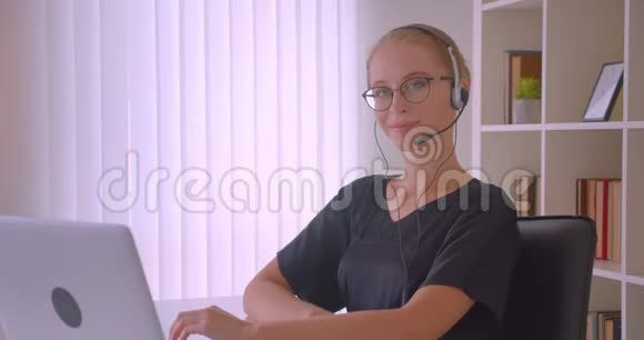 戴着眼镜和耳机的年轻白种人女商人用笔记本电脑看镜头的特写照片视频的预览图