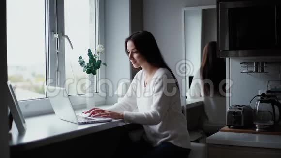 坐在厨房窗户旁边的漂亮女孩在笔记本电脑上打字视频的预览图
