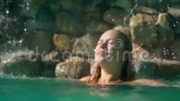 美丽的金发女人在瀑布里放松和享受水滴随着太阳的照射而发光概念放松水疗中心视频的预览图