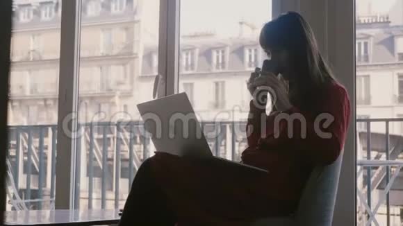 美丽轻松的年轻女商人使用笔记本电脑和智能手机在巴黎的大公寓窗口喝咖啡视频的预览图