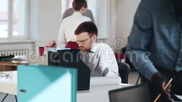 穿着疲惫的年轻英俊的欧洲男性商务经理在繁忙的现代办公桌上使用笔记本电脑视频的预览图