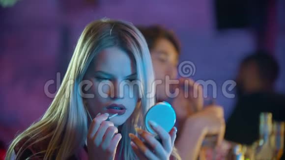 年轻漂亮的金发女郎坐在酒保的旁边站着在嘴唇上涂着红色的口红看着酒保视频的预览图