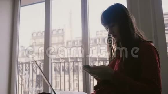 快乐成功的年轻美女博主用笔记本电脑和智能手机在线购物在巴黎的酒店窗口微笑视频的预览图