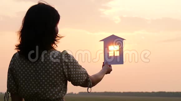 一个穿着连衣裙的女孩在夕阳下拿着纸房子的剪影建设房地产经营视频的预览图