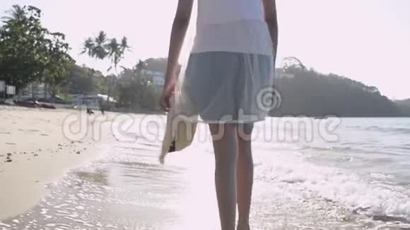 从亚洲女孩背着草帽穿着休闲服装光着脚在阳光下的海滩上行走视频的预览图