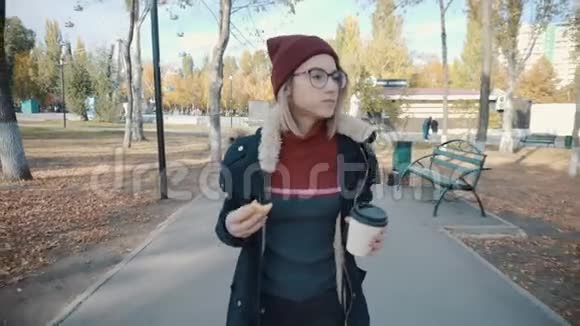 女孩在公园里散步吃汉堡喝咖啡穿着夹克和帽子的女孩视频的预览图