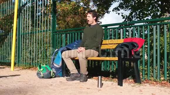 那个家伙坐在城市公园的长凳上打哈欠视频的预览图