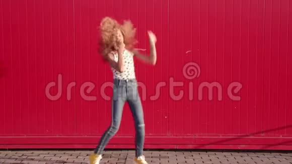 穿着圆点花纹T恤和蓝色牛仔裤跳舞的卷发金发女孩在红墙上闲逛假装潜水视频的预览图