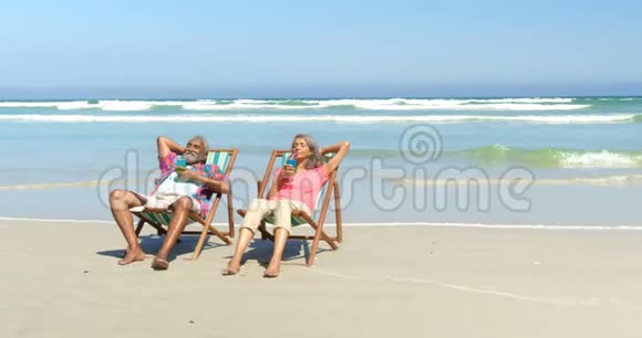 活跃的非裔美国人夫妇在海滩4k的躺椅上喝酒的正面景色视频的预览图