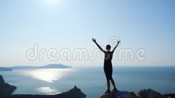年轻受启发的女人举起双手站在海面上一座山的顶端映衬着美丽的蓝天视频的预览图