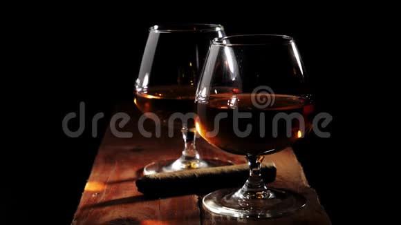 豪华白兰地两杯金色白兰地和黑色背景的棕色木桌上的雪茄手拿雪茄视频的预览图