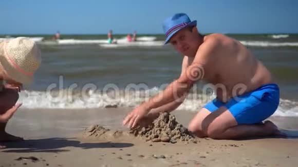 胖爸爸戴着帽子小女儿在潮湿的沙滩上建城堡视频的预览图