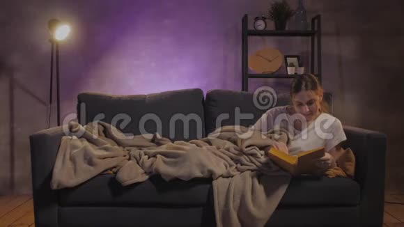 年轻漂亮的女孩躺在沙发上看书女孩看电视视频的预览图