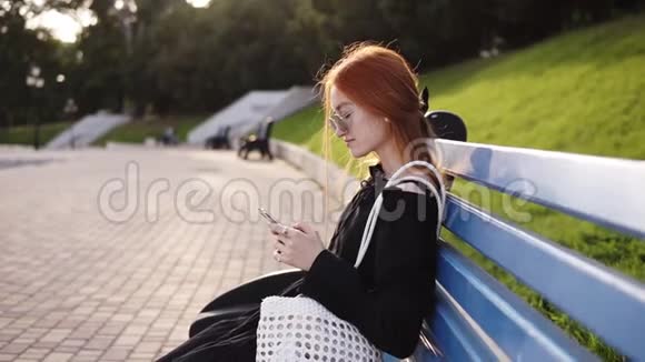 坐在长凳上的城市公园里一个冷静的红头发女孩用智能手机穿着黑色连衣裙和白色袋子的年轻女人视频的预览图