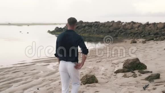 孤独的男人穿着白色的裤子和衬衫沿着沙滩散步背面的景色视频的预览图