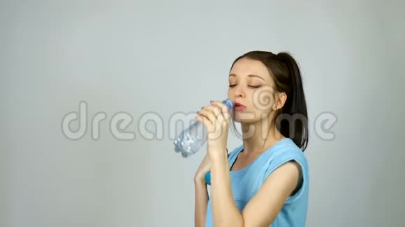 一个长着深色头发的布鲁内特女孩在灰色的环境下锻炼后正在用塑料透明瓶子喝纯净水视频的预览图