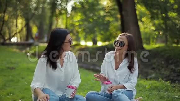 两个穿着白色衬衫的时髦快乐微笑的女孩正坐在公园里绿树成荫视频的预览图