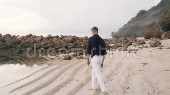 孤独的男人穿着白色的裤子和衬衫沿着沙滩散步背面的景色视频的预览图
