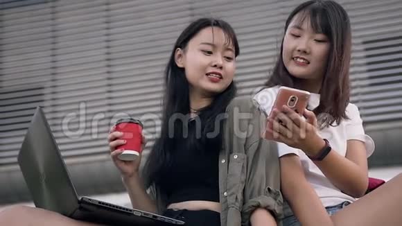 美丽快乐微笑的亚洲女孩坐在大城市大楼附近用电话看照片视频的预览图
