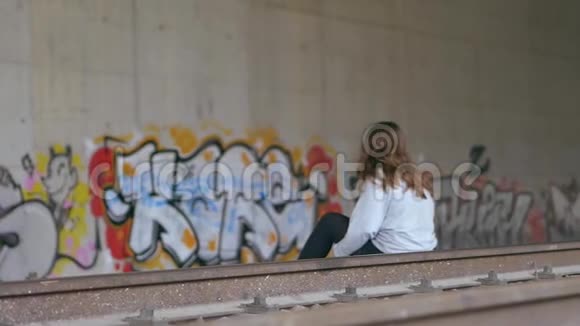 一个长着棕色头发的白人年轻女人坐在火车轨道上隧道墙上的涂鸦慢动作视频的预览图