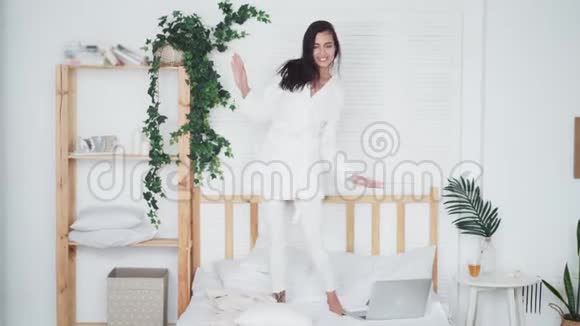穿着白色浴袍带着枕头在床上跳舞的快乐年轻女子视频的预览图