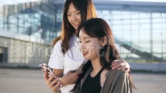 两个活泼可爱的年轻亚洲女孩在现代建筑附近的电话中微笑着修改有趣的照片视频的预览图