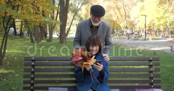 快乐的老夫妻正坐在秋天舒适的城市公园里的长凳上使用手机的男人视频的预览图