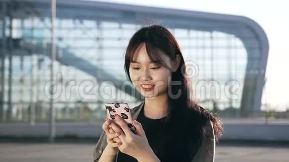 美丽的微笑年轻的亚洲女人走在机场大楼附近用她的手机视频的预览图