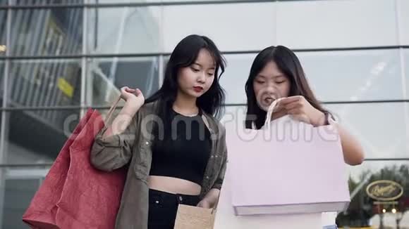两个快乐可爱的年轻亚洲女朋友站在购物中心附近互相展示他们买了什么视频的预览图