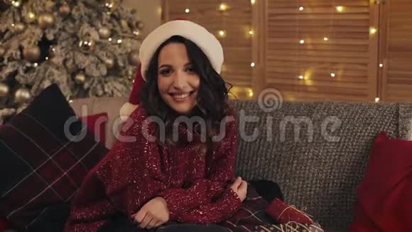 放大年轻美丽的黑发女子戴着桑塔帽坐在圣诞树附近的沙发上闪闪发光的灯光视频的预览图