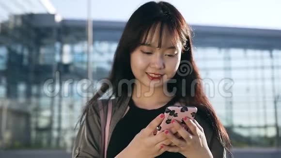 怡然自得的年轻亚洲女性在机场大楼附近的手机上输入短信视频的预览图