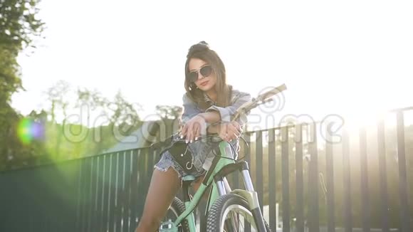 美丽的年轻女子穿着牛仔裤戴着墨镜坐在自行车上对着镜头摆出真诚的微笑视频的预览图