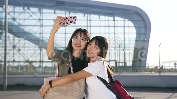 微笑着兴高采烈的亚洲年轻女性穿着现代服装拥抱并在机场附近自拍视频的预览图