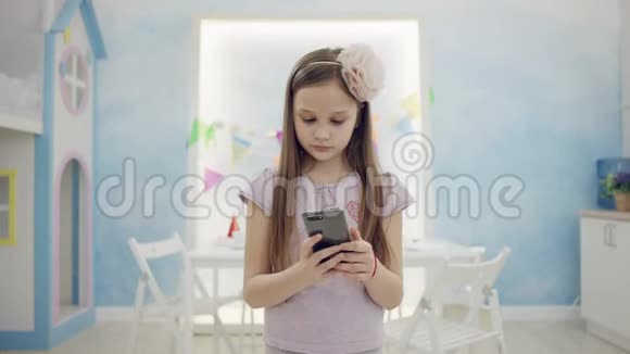 漂亮的小女孩在明亮的房间里站在镜头前用智能手机微笑视频的预览图