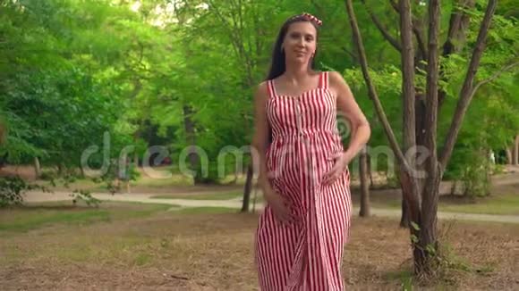 一个怀孕的女孩穿过公园一个穿着条纹白色和红色连衣裙的女孩微笑着跳舞视频的预览图