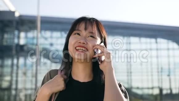 一个年轻的亚洲女人头发乌黑在电话里说话白天在机场大楼附近微笑视频的预览图