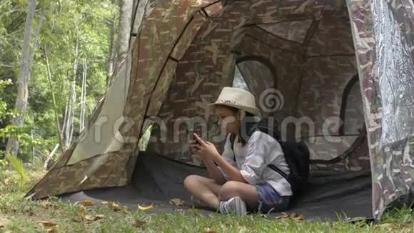亚洲可爱的女孩背包坐着在自然公园的帐篷前使用手机视频的预览图