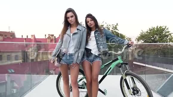 两位穿着时髦牛仔裤衣服骑着自行车和滑板在镜头上摆姿势的白种人年轻迷人的肖像视频的预览图