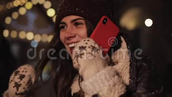 带上戴着冬帽和手套的漂亮女孩在智能手机上说话在坠落中散步时大笑视频的预览图