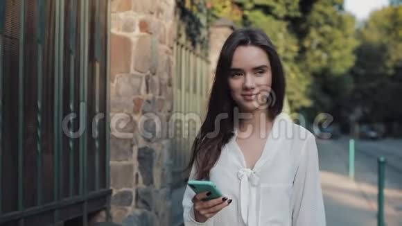 年轻迷人的微笑着的黑发女孩在舒适的街道上一边使用手机一边在社交网络上聊天视频的预览图