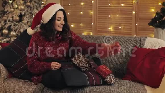 圣诞老人帽里美丽的年轻女子在圣诞礼物上完成了鞠躬仪式并把盒子放在一旁坐在沙发上视频的预览图