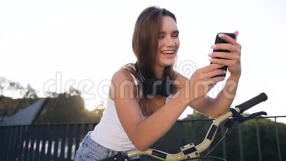 夏天一位留着长发的迷人微笑女孩坐在自行车上在智能手机应用程序中观看一些东西视频的预览图