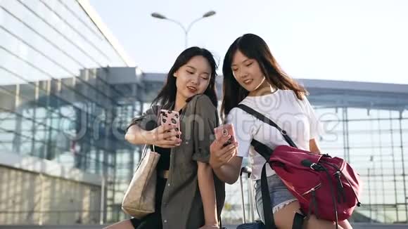 两位迷人精力充沛的亚洲女性坐在行李箱上在大个子附近使用智能手机视频的预览图