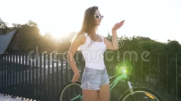 可爱的女孩留着长发戴着时髦的太阳镜穿着诱人的衣服在她的自行车旁的灯光下跳舞视频的预览图