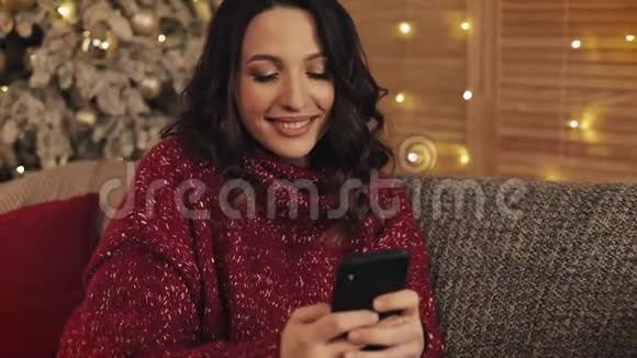 一位年轻漂亮的黑发女人坐在灰色沙发的圣诞树旁用智能手机打字的肖像视频的预览图