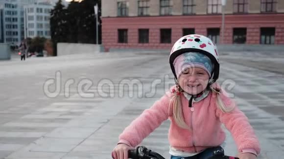 一个穿着粉红色夹克的微笑着的小快乐女孩正骑着绿色自行车在一个城市的夕阳下带着小自行车的视频的预览图