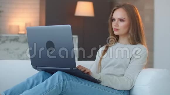 年轻女孩带着笔记本电脑坐在家里的沙发上在沙发上躺着的笔记本电脑键盘上打字沙发在视频的预览图