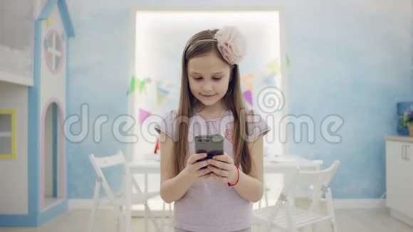 迷人的小女孩在智能手机上玩脸上带着微笑站在镜头前明亮的视频的预览图
